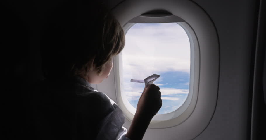 Boy flies in airplane and looks out portholes, holding paper plane toy in his hands. Child tries to cope with boredom of long flight by playing with paper toy. The boy is trying to cope with boredom. Royalty-Free Stock Footage #1100541537