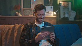 Shot of smiling handsome young Caucasian man sitting on sofa and holding digital tablet indoors. Footage of good-looking unshaven guy enjoying surfing internet in cozy home. Evening - Powered by Shutterstock - Get 15% off with code: PIKWIZARD15