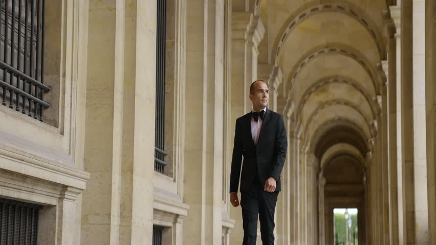Man in black and white official suit walking in arcuate shaped hall. Action. Man walking outdoors by the building with columns.