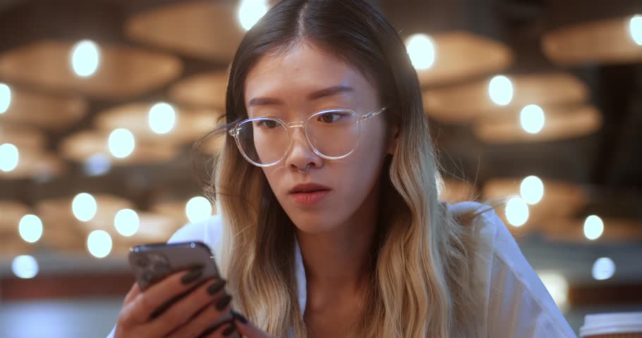 close-up of a young business woman of asian ethnicity uses a mobile phone in the office. Young business woman in glasses uses a mobile phone, scrolling mobile internet, texting message online