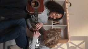 Vertical video. Guitar lesson. Beloved couple. Home learning. Inspired man showing woman accords on acoustic instrument sitting light room interior.