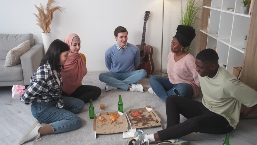 Present exchange. Friends party. Holiday surprise. Positive men and women sitting floor with pizza showing each other gift boxes in light room interior.
