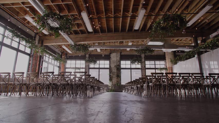 Wedding ceremony decor florals and candles set up in industrial warehouse event space; greenery chandeliers hanging from ceiling | Shutterstock HD Video #1100558973