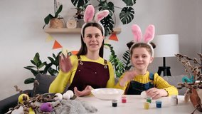 Pretty little daughter and smiling young mom in fluffy bunny ears talk on video call, looking at camera, funny mom and cute little girl child sitting at table in decorated room. Happy easter concept