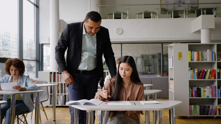 Positive African American male teacher at high school institution, wearing eyeglasses and formal suit, helping a female Asian student, pointing at textbook while explaining lesson in the classroom Royalty-Free Stock Footage #1100568137