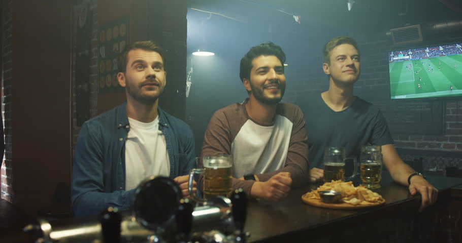 Three handsome men standing at the pub in the evening and cheering up while watching sport game. Cheerful sport fans. | Shutterstock HD Video #1100568309