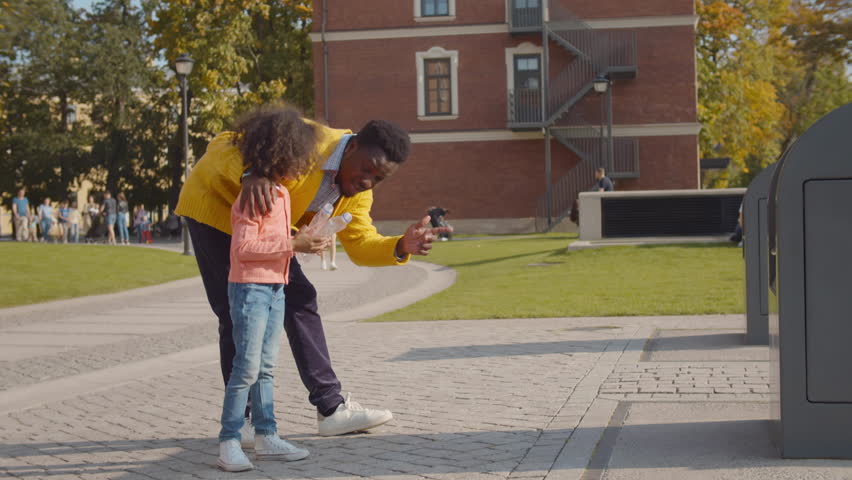 Father and little daughter throw empty bottle into recycling bin. African-American dad and child throw waste in city container. Family throwing waste in bin. Environment, ecology concept. Realtime Royalty-Free Stock Footage #1100568637