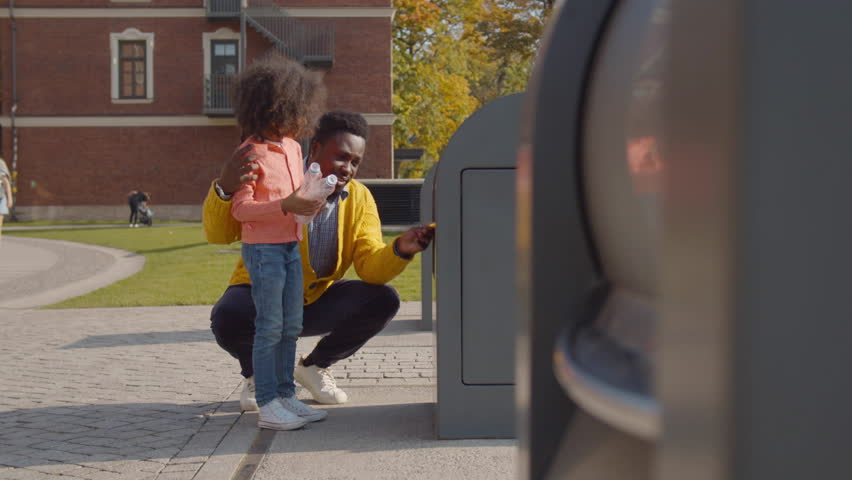 Father and little daughter throw empty bottle into recycling bin. African-American dad and child throw waste in city container. Family throwing waste in bin. Environment, ecology concept. Realtime | Shutterstock HD Video #1100568637