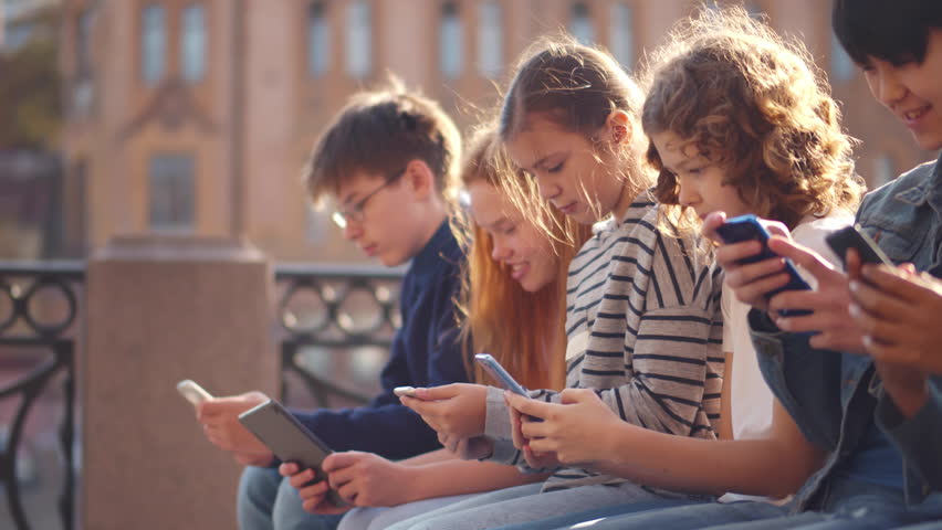 Multiethnic group of school students outdoors use mobile phones. Kids sit in row and play on cellphones outdoors. Diverse kids use smartphone in city. Realtime Royalty-Free Stock Footage #1100568641