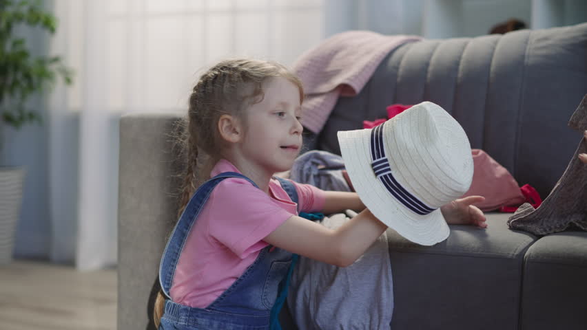 Little girl puts stylish straw hat into suitcase with clothes on sofa closeup. Child helps gather items for vacation. Accessories for rest on beach | Shutterstock HD Video #1100577169