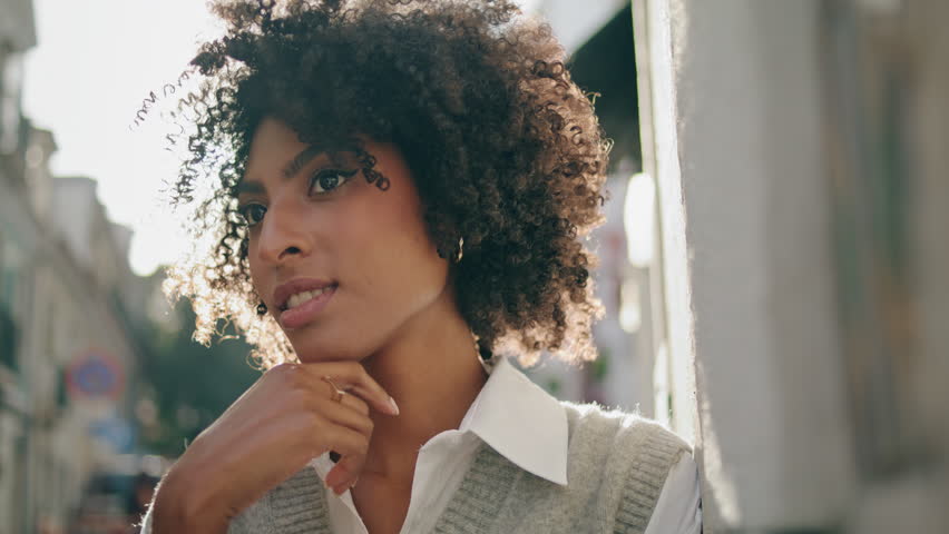 Cute attractive lady relaxing dreaming on sunny city street close up. Happy african american woman standing outdoors under bright sunlight looking camera. Portrait of beautiful curly brunette in town. | Shutterstock HD Video #1100580645