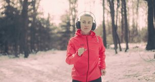 Running woman, headphone girl runner on the snow in winter sunny day. Female in pink sportswear jacket fitness training outdoors. Sport concept, 4K slow motion video
