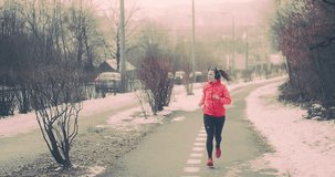 Running woman, headphone girl runner on the snow in winter sunny day. Female in pink sportswear jacket fitness training outdoors. Sport concept, 4K slow motion video
