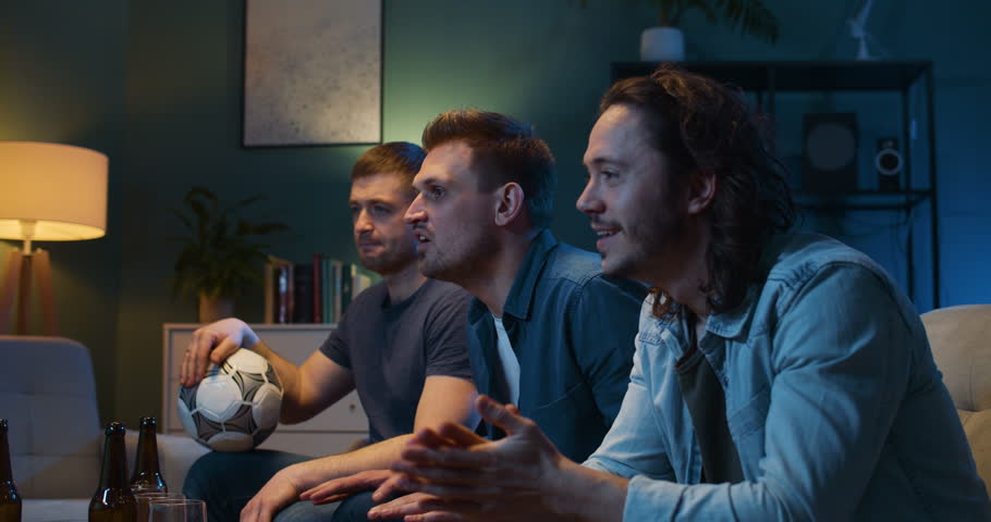 Tensed Caucasian male football fans screaming, worrying and cheering for favorite team while watching TV and sitting late at night in living room. Guys friends having nice evening together at home. | Shutterstock HD Video #1100583847