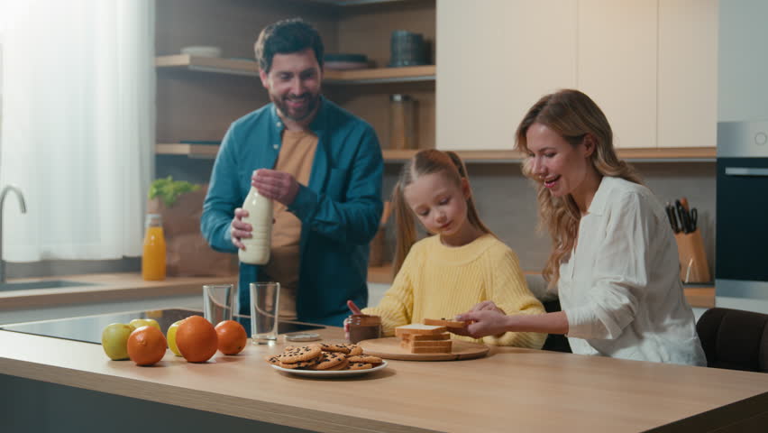 Caucasian family cooking breakfast together at kitchen at home mother with little daughter child girl prepare toasts bread with chocolate pasta father pour milk into glasses parents with kid cook Royalty-Free Stock Footage #1100584723