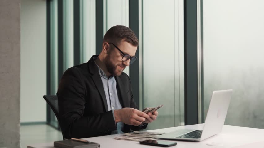 A happy young man in business clothes counts money bills while sitting at a laptop Royalty-Free Stock Footage #1100586585
