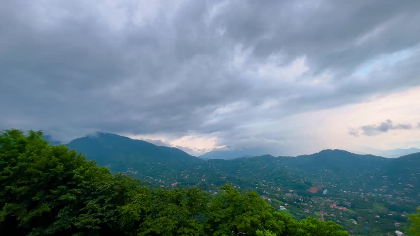Panning shot: view from observation deck to mountains with green trees, rural houses, grey cloudy sky and city of Batumi in the distance in the afternoon: slow motion. Georgian landscape in Georgia