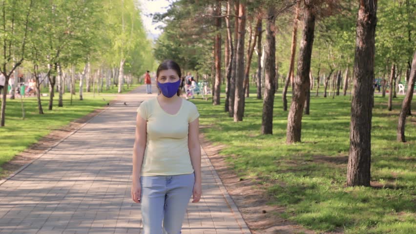 A young woman in a blue mask on her face is walking in the park in the evening.