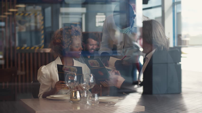 Waiter making recommendation for diverse couple in a restaurant. View through window of multiethnic couple order dinner in cafe. Waiter in mask take order from couple customers. Realtime | Shutterstock HD Video #1100603389