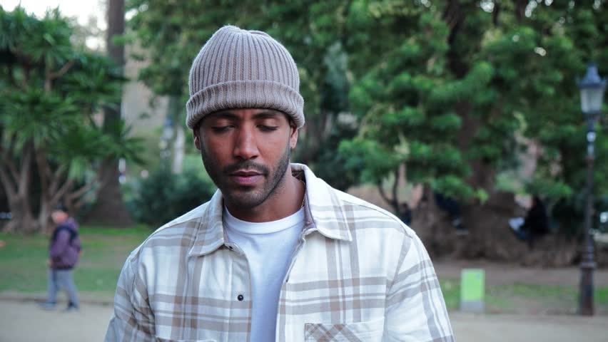 Close up portrait of young attractive african american man with beanie hat looking serious at camera. Front view of a hispanic guy standing in a park outdoors with sad attitude. Slow motion. High