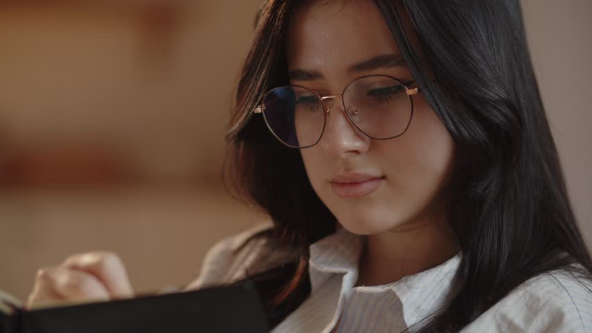 A close-up shot of a woman in glasses writing in notebook. A cute girl holds a pen in her hands and makes notes. Charming brunette student working on essay