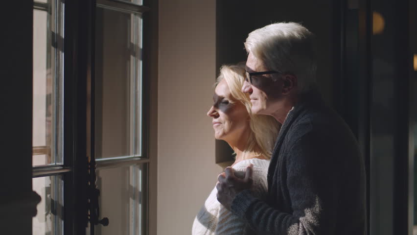 Happy senior couple embracing and looking through window together. Romantic aged man and woman hug standing near window. Active retirement and lifestyle concept. Realtime | Shutterstock HD Video #1100611111
