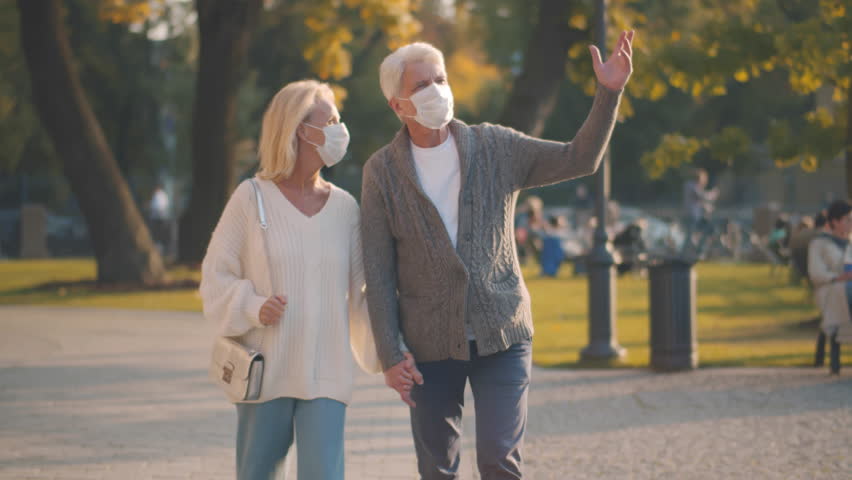 Portrait senior couple in medical mask walk talking in autumn park during covid-19 pandemic. . Mature man and woman in protective mask walk outdoors. Aged couple in mask in park. Realtime | Shutterstock HD Video #1100611185