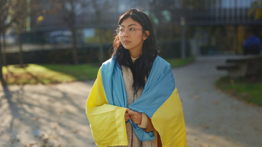 Portrait of the pretty Asian woman is smiling and wrapped in the Ukrainian flag looking at the camera. Glory to Ukraine. Patriot. Stand with Ukraine. Support