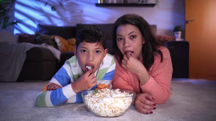 Cheerful mother and son spending time together at home in the nighttime, watching a movie lying on the floor in the living room and eating popcorns. Point of view of latin family looking at camera. | Shutterstock HD Video #1100612419