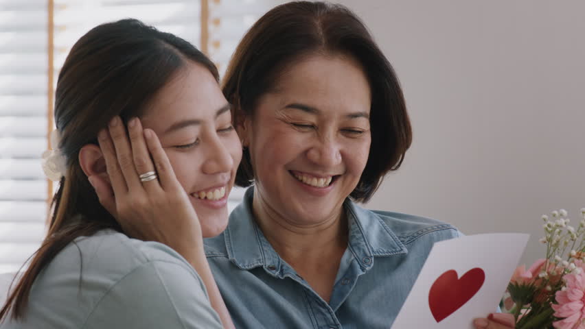 May Mother's day young adult grown up child cuddle hug give flower gift box red heart card to mature middle aged mum. Love kiss care mom asia people sitting at home sofa happy smile enjoy family time. Royalty-Free Stock Footage #1100613281