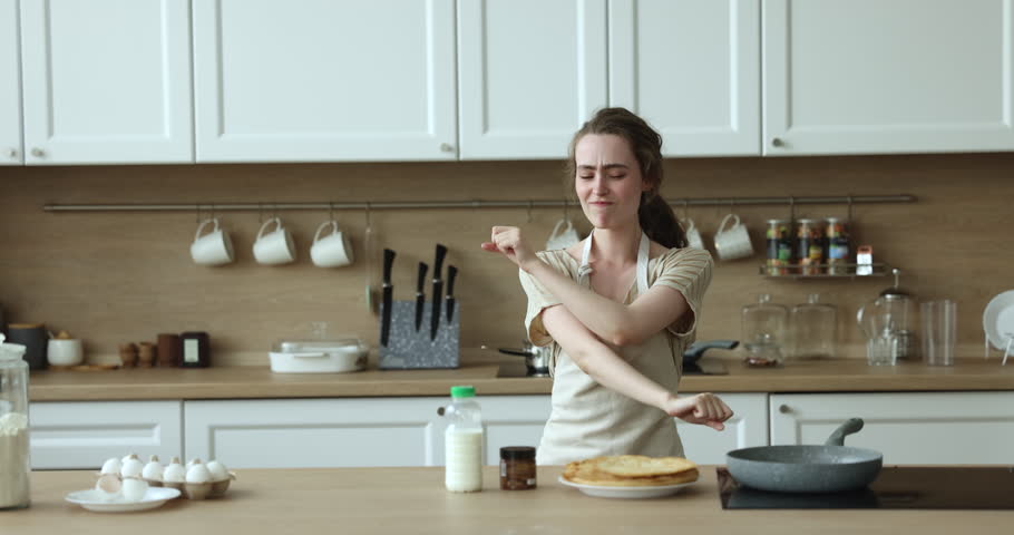 Young 25s cheerful woman in apron moves to favourite music, dancing feels happy, looks carefree while cooking homemade pancakes in modern kitchen, spend leisure prepare breakfast enjoy fun and cookery Royalty-Free Stock Footage #1100614333