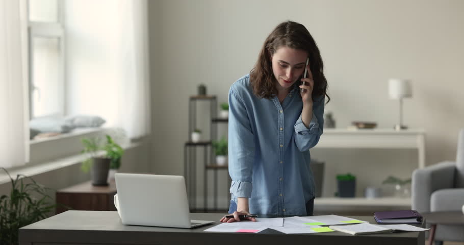 Young busy woman working on project, takes notes on paper, prepare report, doing research and talking to company client or colleague on cellphone, having business call at workplace, working in office Royalty-Free Stock Footage #1100614343