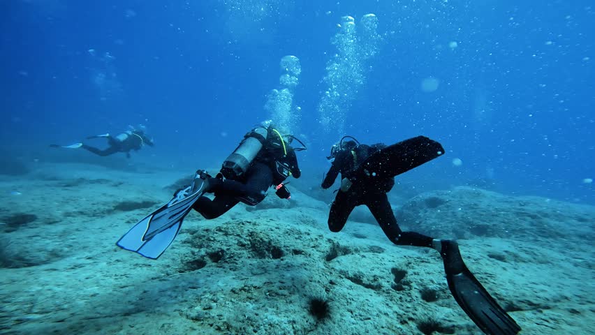 Two men scuba diving relaxed underwater follow shot; breathing and kicking with fins; deep blue aegean sea; swimming over rocks and sand on bottom of ocean; calm beautiful aquatic adventure Royalty-Free Stock Footage #1100619231