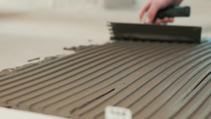 Spreading tiles adhesive on a floor with notched trowel. Construction worker placing tiles.