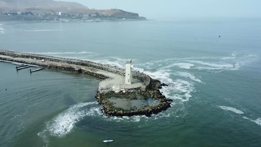 Drone orbits around a white small lighthouse in the edge of a rocky pier. In the background coastal mountain begin to appear as the camera turns left. Located in Lima, Peru.