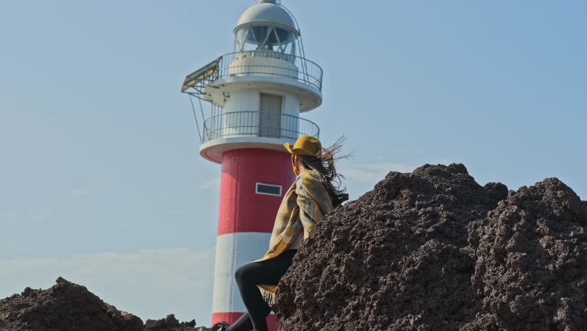 Traveling Woman On A Windy Day In Tenerife, Spain, Leaning On Rock With Punta de Teno Lighthouse In Backdrop. low angle, slowmo