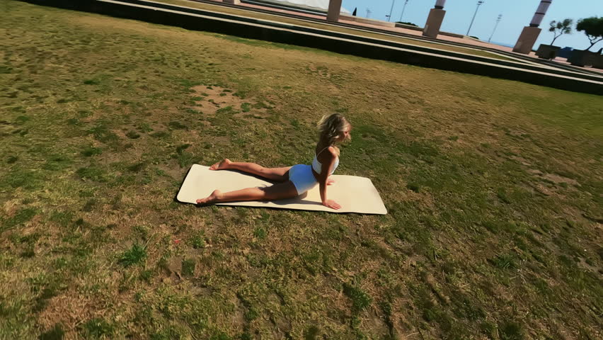 Dynamic fpv drone shot flying around girl in white dress doing stretching exercises on the grass at sunny day and using mat. High quality 4k footage