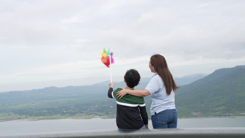 A progressive woman and her son are on vacation, enjoying the natural beauty of a lake at the bottom of a hill while the boy carries a toy windmill.