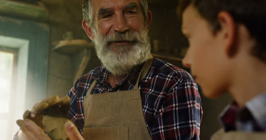 Close up of the old Caucasian grandpa and cute little grandson sitting together in the pottery manufacture and having their hands in clay.
