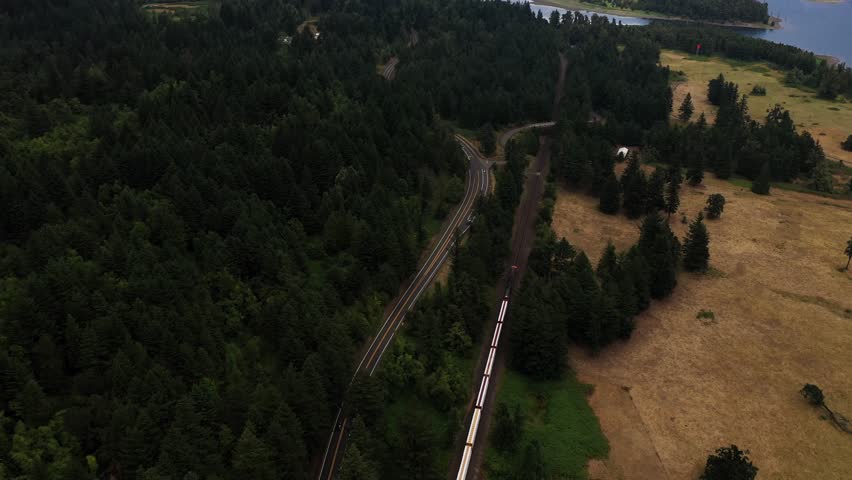 Aerial view of train driving on tracks along river bank of the Columbia River Gorge in Washington and Oregon surrounded by forest of pine trees blue river and highway