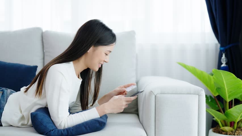Young asian woman lying on sofa in living room, makes online banking payments through the internet from bank card on smartphone. Shopping online on mobile phone with credit card | Shutterstock HD Video #1100638669