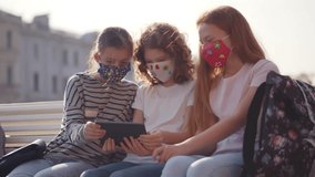 Schoolgirls in mask use smartphone together outdoors. Group of teen female friends watch video on digital tablet in city. High school students with tablet pc wear medical mask. Realtime. 