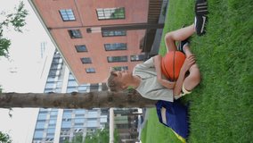 Caucasian boy sitting in public city park with baskaetball ball and looking at camera. Vertical footage.