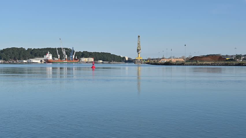 Interior Mercantile Port of Bayonne, France, in the foreground the calm water of the river flows, in the background a maroon ship with three white cranes in the middle a red buoy,a large yellow crane