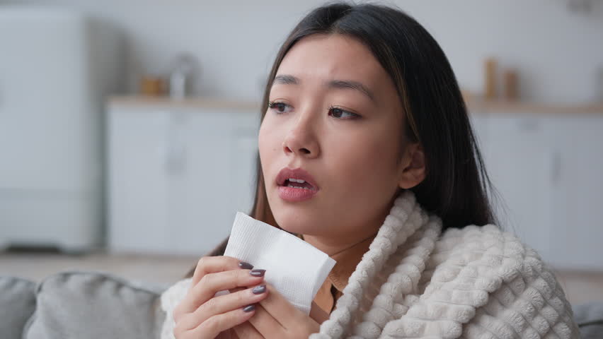 Sick Asian woman covered in blanket sneezing in paper napkin sneeze runny nose feel frozen cold unhealthy korean girl patient has covid symptom grippe nasal infectious disease health treatment at home Royalty-Free Stock Footage #1100655759