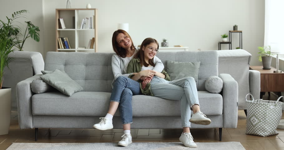 Smile adult daughter relax on sofa with dear mom feel safe in warm embrace her beloved mother. Loving parent sits on couch, hugs caressing to teen child. Attractive happy family enjoy weekend at home | Shutterstock HD Video #1100656527