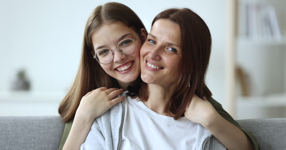 Teenage daughter embrace mother from behind and look at camera, close up portrait of beautiful middle-aged woman enjoy cuddles spend time together with pretty teen child in glasses. Family ties, love Royalty-Free Stock Footage #1100656549