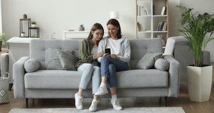 Middle-aged mother and teenager daughter relax on sofa at home, spend leisure look at mobile phone screen, watch video blogs, on-line content. Nice time with family use modern wireless technologies