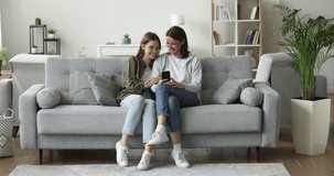 Middle-aged mother and teenager daughter relax on sofa at home, spend leisure look at mobile phone screen, watch video blogs, on-line content. Nice time with family use modern wireless technologies
