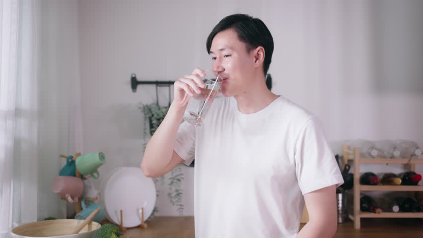 20s young Asian man in white t-shirt drinking a glass of fresh water in kitchen at home. Hydration and hygiene concept Royalty-Free Stock Footage #1100658083